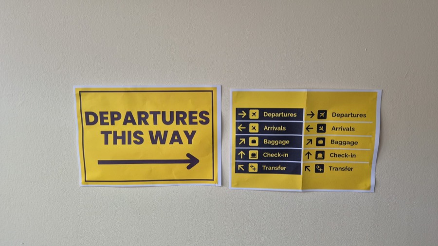 A hand made yellow sign reading Departures This Way, taped to a wall beside a printed list of gates